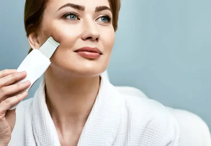 Kobieta w białym szlafroku używa urządzenia do peelingu kawitacyjnego. Dowiedz się, jak używać peeling kawitacyjny dla promiennej cery.