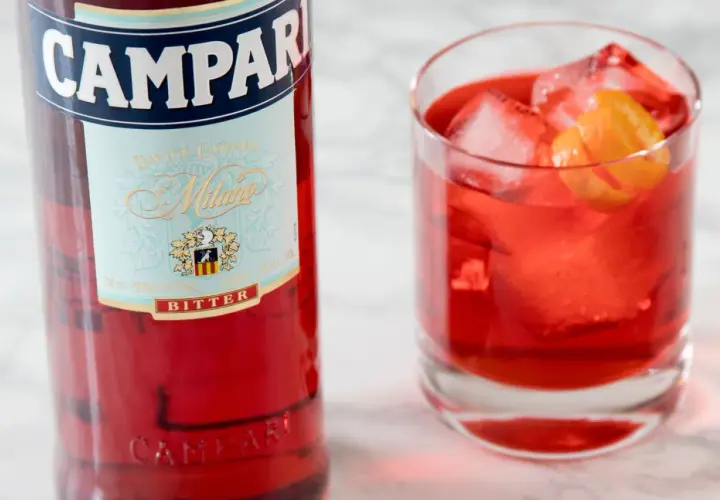 Butelka Campari i szklanka z czerwonym drinkiem, lodem i skórką pomarańczy. Idealny aperitif, który może być bazą dla drinków z Aperolem.