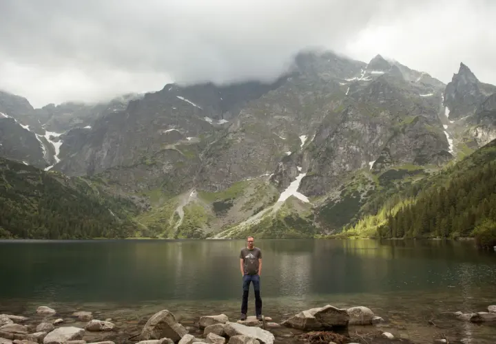Mężczyzna stoi na kamieniach nad brzegiem jeziora Morskie Oko. W tle majestatyczne góry Tatr.