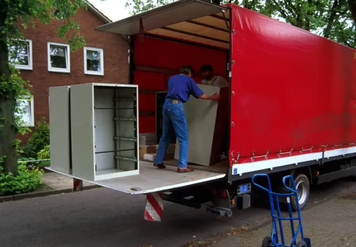 Umzugshelfer laden einen Schrank auf einen roten LKW. Der Schrank steht am Straßenrand, was die Bedeutung von "kerbside" verdeutlicht.