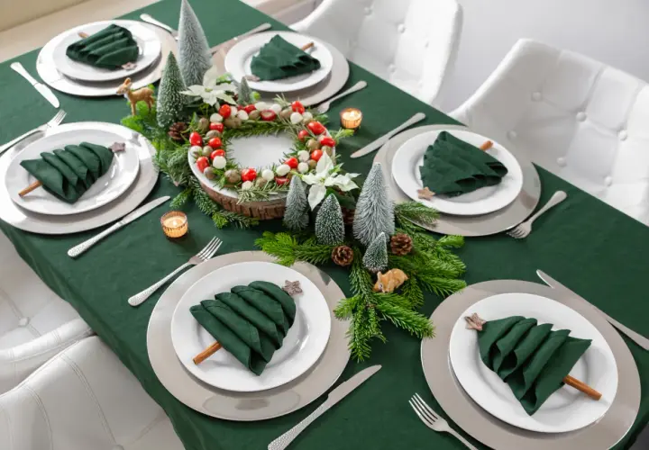Moderne tischdeko weihnachten: grüne Servietten als Bäume, Tannenzweige, Kerzen und ein Kranz mit Beeren auf einem grünen Tischtuch.