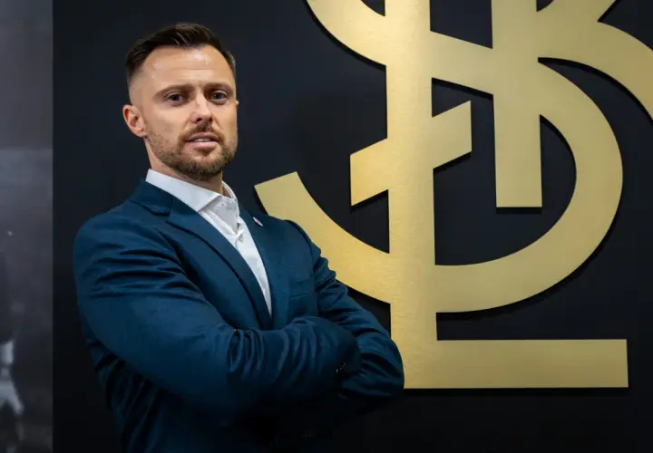 Mężczyzna w garniturze, trener ŁKS Łódź, stoi przed złotym logo klubu.
