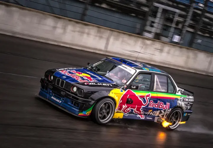 BMW E30 w barwach Red Bull, auto do driftu, z płomieniami z wydechu, na mokrym torze.