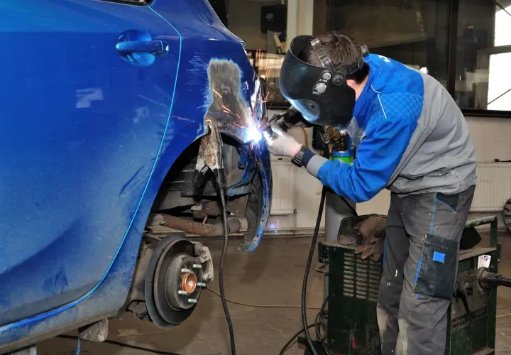 Mechanik w masce spawa blachę samochodową, przywracając jej dawny kształt. Iskry sypią się podczas prac blacharskich.