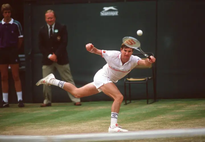 Jimmy Connors w akcji, uderza piłkę na trawiastym korcie. W tle sędzia i chłopiec podający piłki.