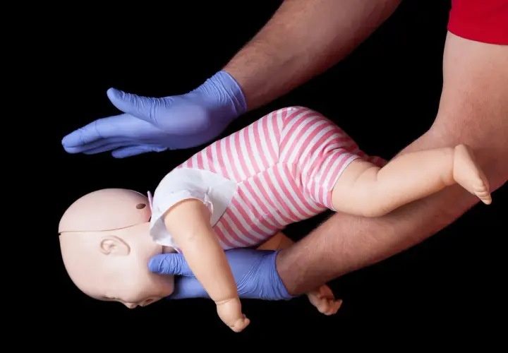 Esercitazione sulla manovra di Heimlich per bambini: mani guantate sostengono un manichino neonato, simulando un intervento di primo soccorso.