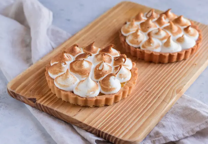 Due crostatine con crema al limone e meringa all'italiana, dorata e soffice, su un tagliere di legno.