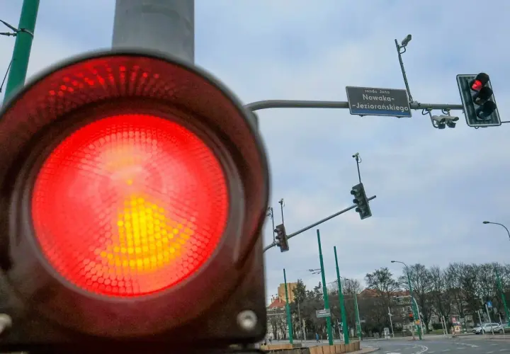 Zbliżenie na czerwone światło sygnalizatora, obok widać rejestrator przejazdu na czerwonym świetle i znak ronda Jana Nowaka-Jeziorańskiego.