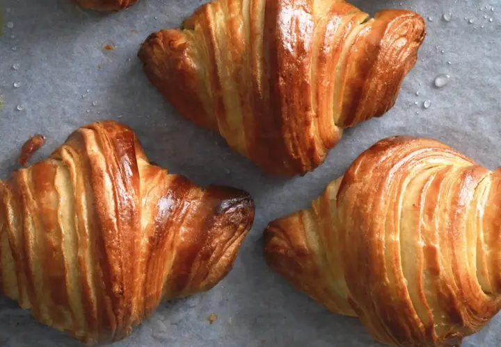Croissant dorati e sfogliati, perfetti per una ricetta croissant fatta in casa. Si intravedono gocce d'acqua sulla carta da forno.