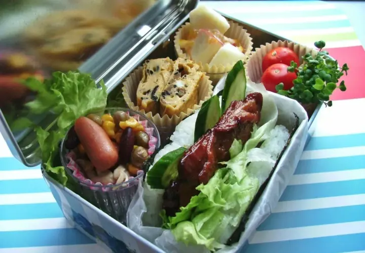 A delicious bento box with a glazed meat skewer, rice, lettuce, egg roll, sausage, beans, corn, tomatoes, and sprouts.