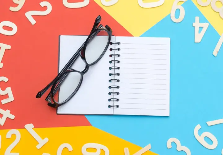 Occhiali neri su un quaderno aperto, circondati da numeri in legno sparsi su sfondi colorati. Un promemoria visivo per imparare l'inglese, magari il numero 66.