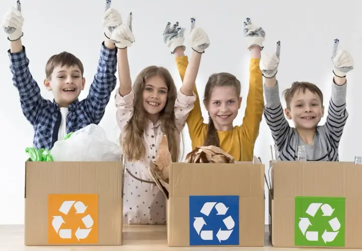 Niños felices participan en un taller de reciclaje, con guantes y pulgares arriba, junto a cajas de cartón con símbolos de reciclaje.
