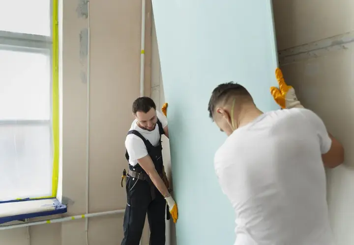 Dwóch budowlańców montuje płytę gipsowo-kartonową, przygotowując ścianę do dalszych prac. Dbałość o idealną spoina jest kluczowa.
