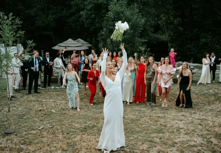 Panna młoda w białej sukni rzuca bukietem, a zgromadzeni goście, w tym panie w kolorowych kreacjach, czekają na jego złapanie. To piękny moment celebrujący tradycje weselne.