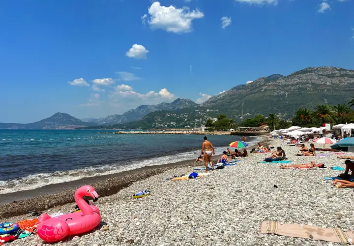 Słoneczny dzień na plaży w Barze, Czarnogóra. Ludzie odpoczywają na kamienistej plaży, a w tle widać góry. Różowy flaming unosi się na wodzie.