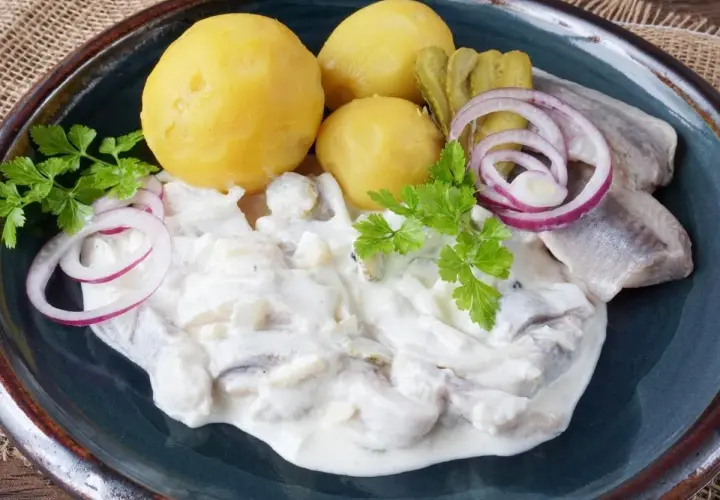 Marinierter Hering in cremiger Sauce mit Salzkartoffeln, Gurken und Zwiebelringen garniert.