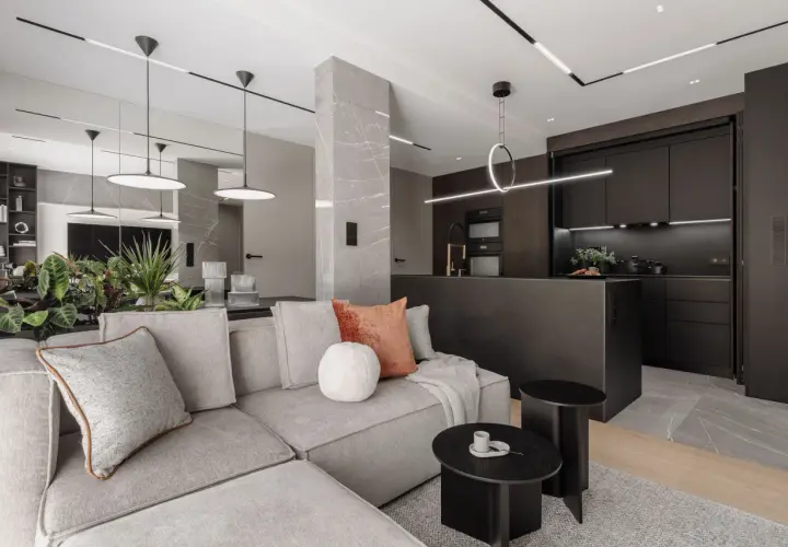 Nowoczesny salon z kuchnią, gdzie oświetlenie wpuszczane tworzy elegancką atmosferę. Szara sofa, czarne meble kuchenne i marmurowe detale.