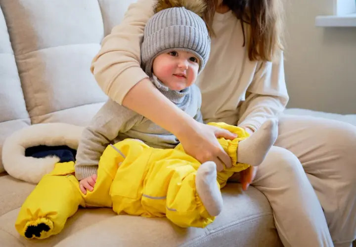 Mama ubiera dziecko w żółty kombinezon i czapkę. Wskazówki, jak ubrać dziecko odpowiednio do temperatury, pomogą Ci przygotować malucha na spacer.