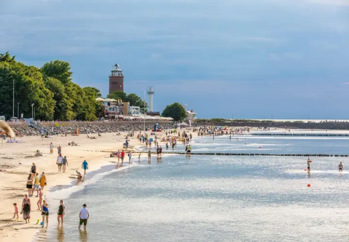 Tłumy ludzi na piaszczystej plaży, latarnia morska w tle. Idealne miejsce na tanie miejscowości nad morzem.