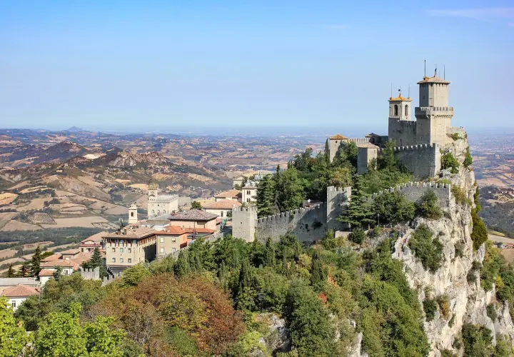 Widok na twierdze i miasto San Marino na wzgórzu, z pagórkowatym krajobrazem w tle.