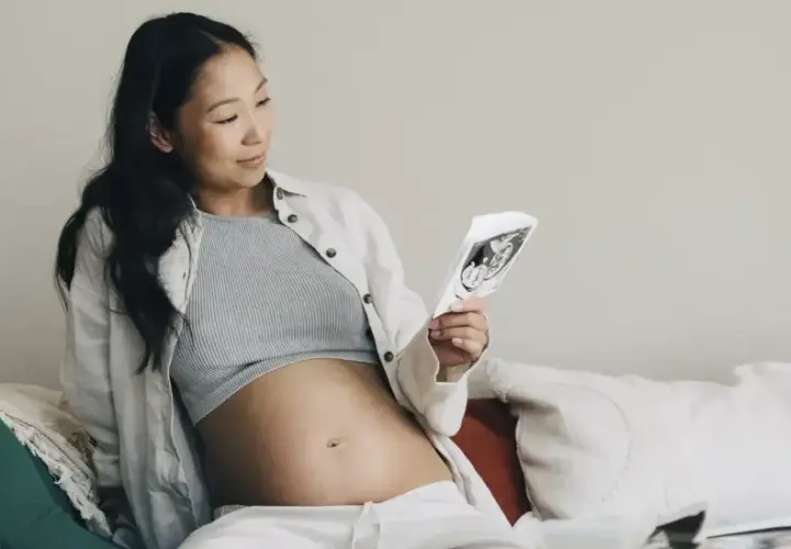 En gravid kvinna i andra trimestern sitter på en soffa och tittar på ett ultraljudsfoto av sitt barn.
