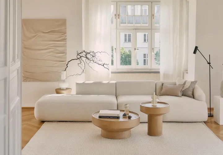Jasny, przytulny salon z beżowym domem w tle. Miękka sofa, drewniane stoliki i subtelne dekoracje tworzą spokojną atmosferę.