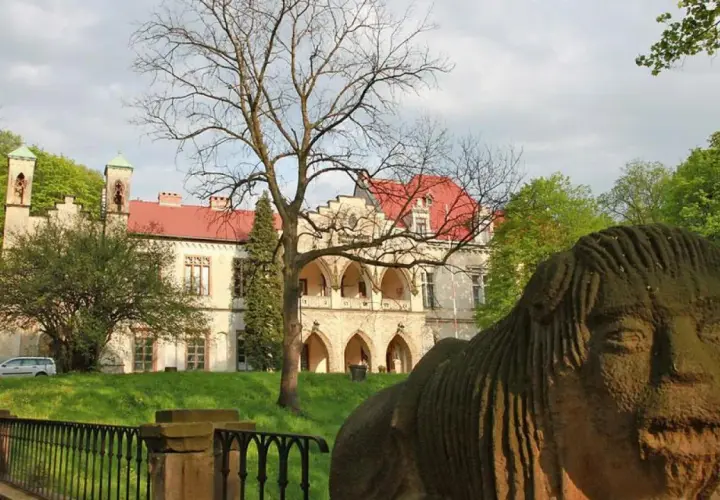 Kamienna rzeźba twarzy przed pałacem w Młoszowej. Budynek z czerwonym dachem i arkadowym gankiem otoczony zielenią.