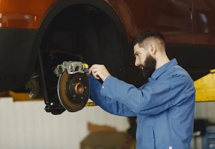 Mechanik w niebieskim kombinezonie pracuje przy hamulcach samochodu. Widać tarczę hamulcową i zacisk. To ważny punkt, od dot 3 a dot 4 zależy bezpieczeństwo jazdy.