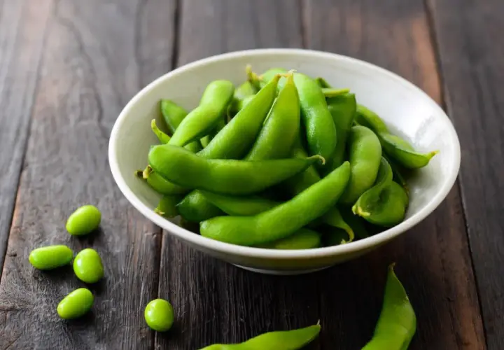 Miseczka pełna zielonej fasolki edamame na drewnianym tle. Kilka fasolek rozsypanych obok.