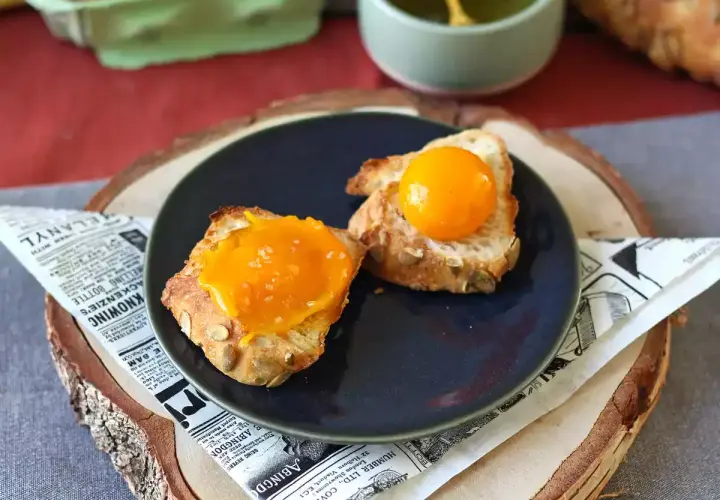 Sur une assiette, deux toasts de pain aux graines, l'un garni d'une texture orangée et l'autre d'un jaune d'œuf confit brillant.