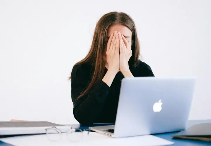 Mujer frustrada cubre su rostro, frente a un portátil. ¿Qué es un falso autónomo? Ejemplos de precariedad laboral.