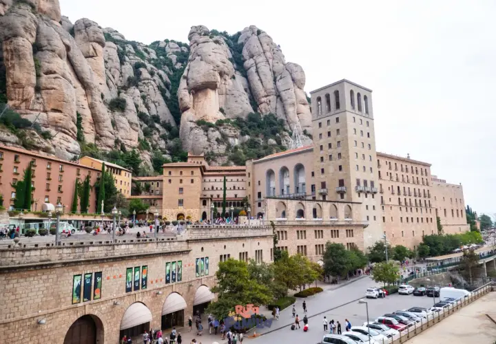 Edificios del monasterio se alzan bajo las imponentes rocas del parc natural de la muntanya de Montserrat. Turistas pasean por la plaza.
