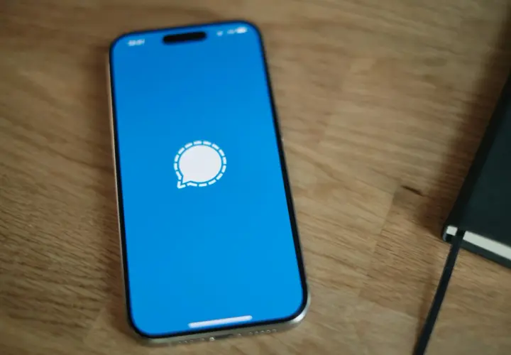 Telefon z niebieskim ekranem i ikoną komunikatora Signal. Obok leży czarny notatnik.