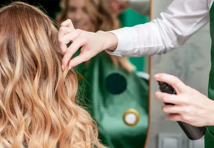 Fryzjerka utrwala fryzurę klientki, spryskując jej blond włosy lakierem. Perfekcyjny styling włosów.