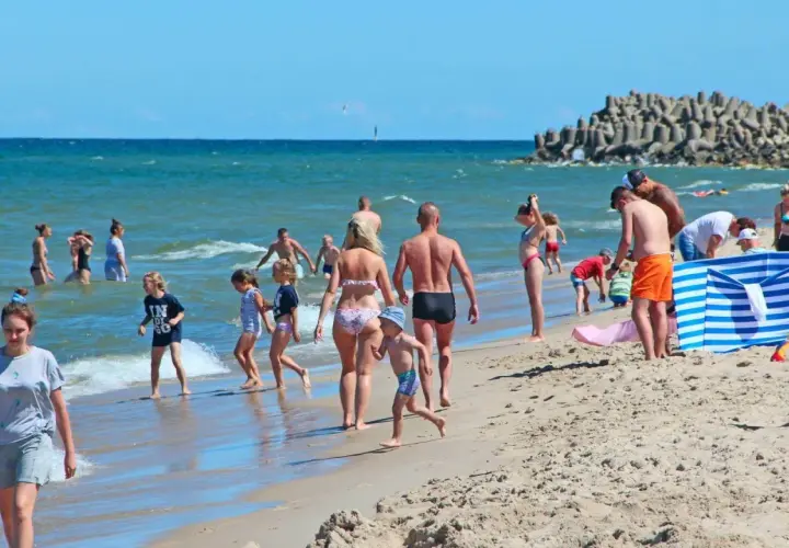 Rodziny cieszą się słońcem i morzem. Widać plażowiczów, dzieci bawiące się w wodzie. Ceny nad morzem w wakacje są wysokie, ale warto.