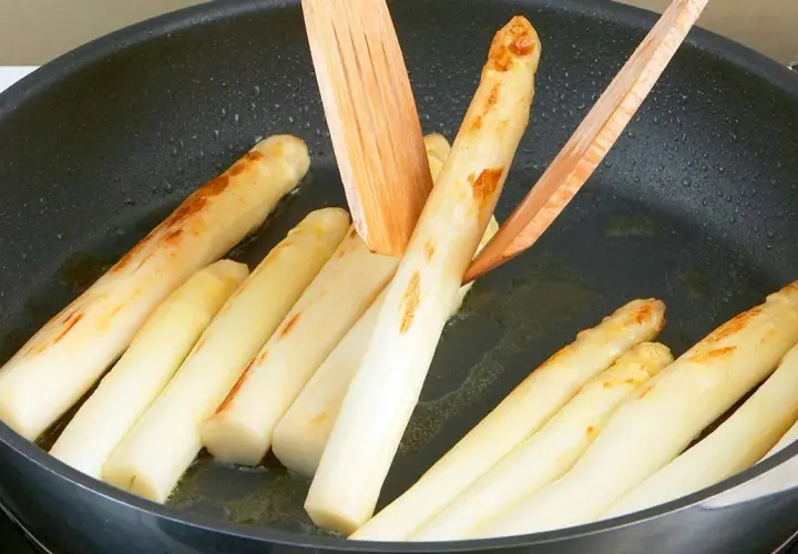 Weißer Spargel brät in einer Pfanne mit etwas Öl. Holzspieße wenden die Stangen, die leicht gebräunte Stellen zeigen.