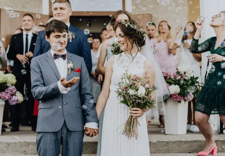 Para młoda trzymająca się za ręce, otoczona bańkami mydlanymi i gośćmi weselnymi.