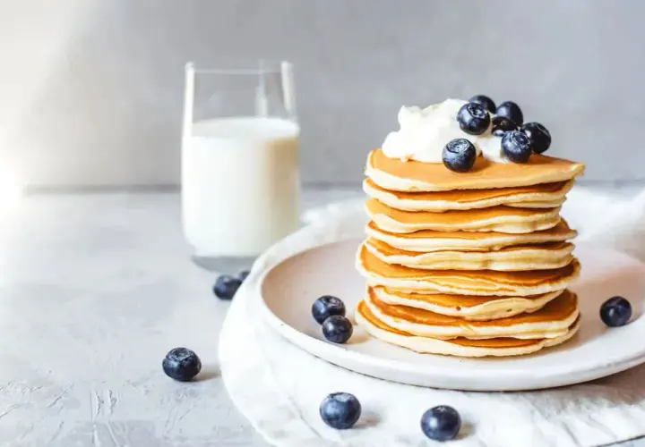 Ein Stapel goldbrauner Protein Pfannkuchen, garniert mit Sahne und Blaubeeren, daneben ein Glas Milch.