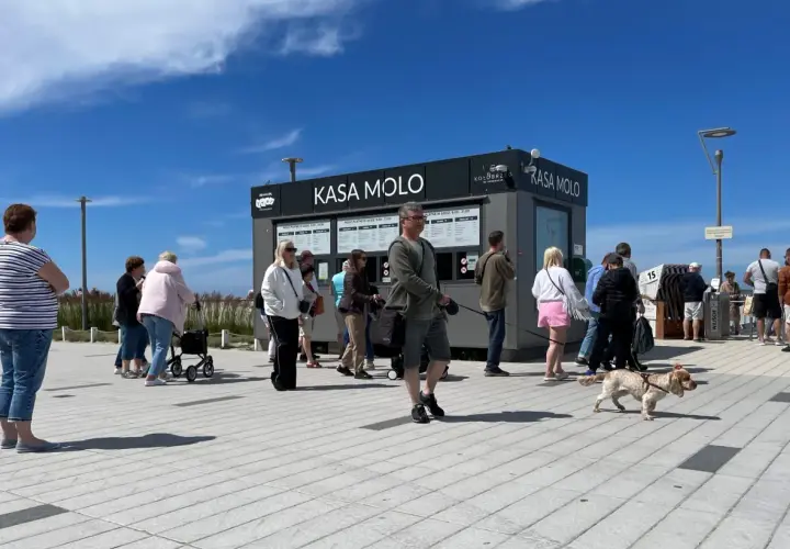 Ludzie stoją w kolejce do kasy na molo w Kołobrzegu. W tle czarny budynek z napisem "KASA MOLO".