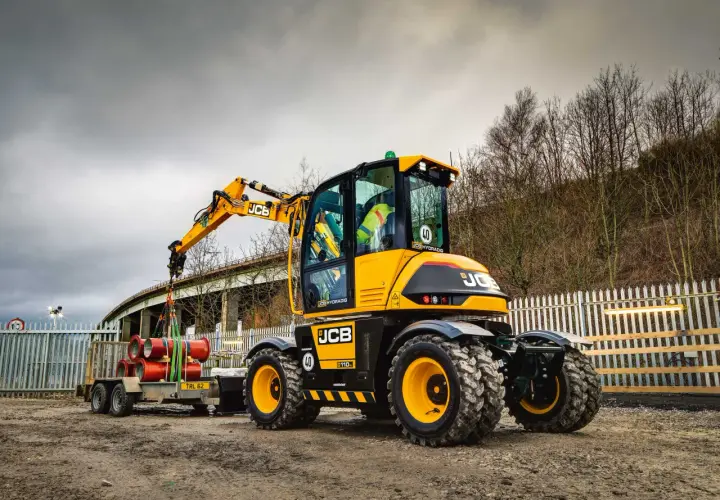 Żółta koparka JCB Hydradig, ważąca ok. 11 ton, ładuje rury na przyczepę.