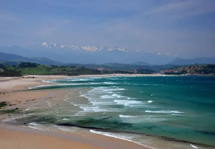 Amplias playas de arena dorada y aguas turquesas, con olas rompiendo suavemente. Al fondo, colinas verdes y montañas nevadas, un paisaje idílico de playas cerca de Comillas.