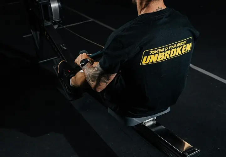 Mężczyzna z tatuażami na ramieniu trenuje na wioślarzu. Czy to crossfit czy siłownia, ważne, że jest UNBROKEN!
