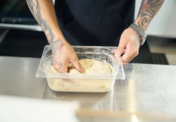 Ręce z tatuażami zagniatają ciasto na pizzę w pojemniku. Czy można mrozic ciasto na pizze? Tak, to dobry sposób na przechowywanie.