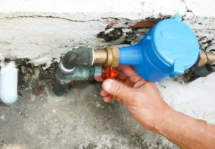 Main d'une personne actionnant le robinet rouge du tuyau d'arrivée d'eau de la maison, juste avant le compteur bleu.