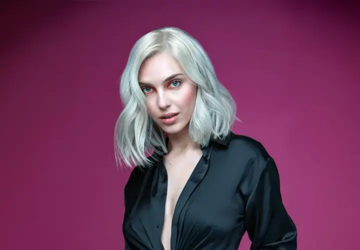 Mujer con cabello rubio ceniza platino, ojos azules y camisa negra abierta, sobre fondo rosa.