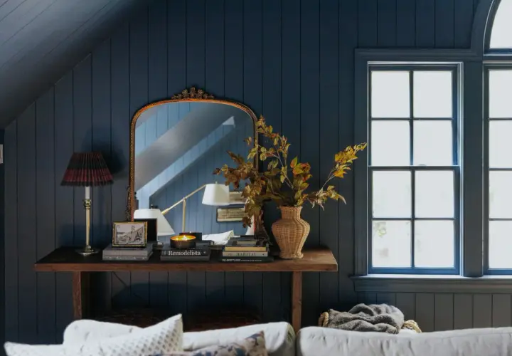 A rustic style console table with a lamp, books, and a vase of autumn leaves sits against a dark blue paneled wall, reflecting in an ornate mirror.