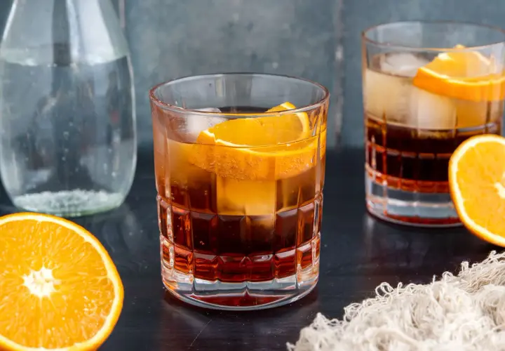 Ein erfrischender Americano Cocktail mit Eis und Orangenscheiben, daneben eine Karaffe Wasser und frische Orangen.