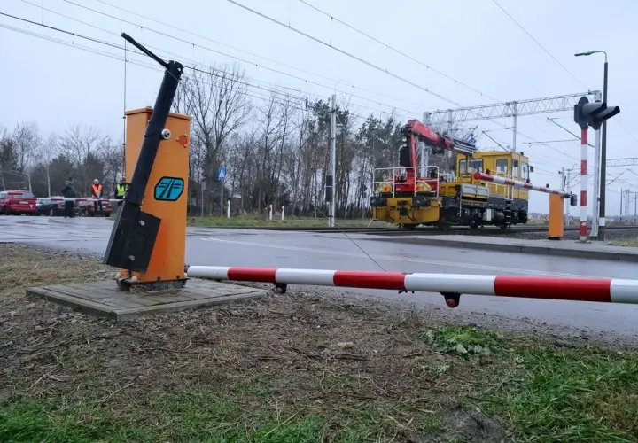 Żółta lokomotywa z dźwigiem zbliża się do przejazdu kolejowego bez zapór. Na pierwszym planie pomarańczowy słup z podniesionym ramieniem.