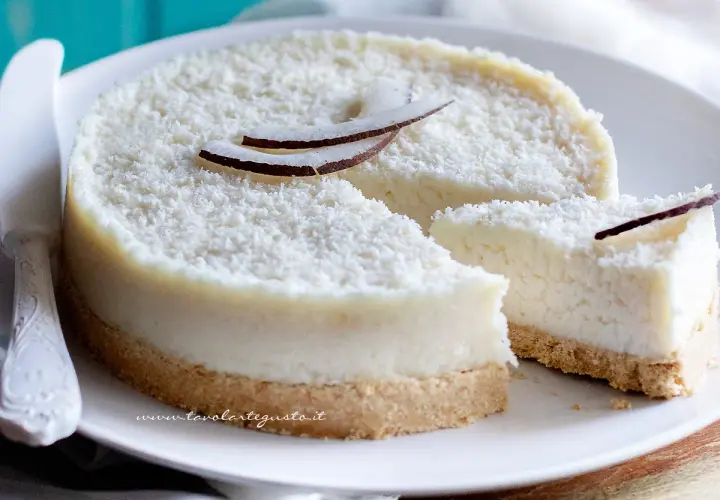 Delizioso cheesecake al cocco, ricoperto di cocco grattugiato e decorato con scaglie di cocco fresco. Una fetta è già stata tagliata, invitando a gustare questo dolce tropicale.