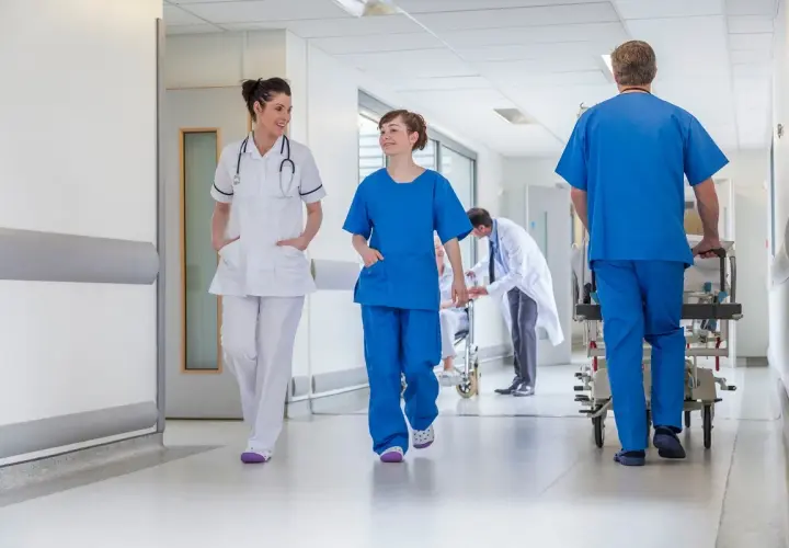 In un corridoio ospedaliero, personale medico si muove con urgenza. Un uomo spinge una barella, mentre due infermiere camminano velocemente, forse a seguito di un evento avverso.